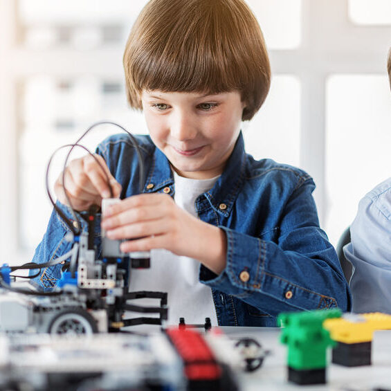 Cheerful boy is making small robot and looking at it with curious smile. He sitting beside friend in workshop